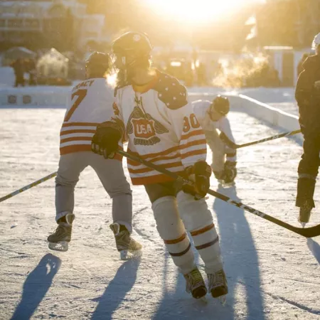A hockey player in Team USA gear skates on an outdoor ice rink, with teammates and shadows stretching in bright sunlight, too.