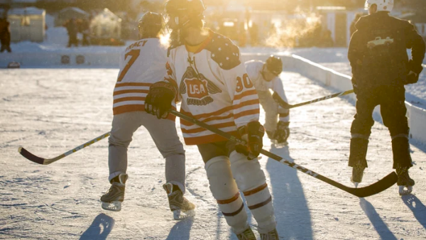 A hockey player in Team USA gear skates on an outdoor ice rink, with teammates and shadows stretching in bright sunlight, too.