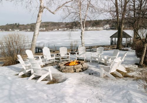 Snowy lakeside patio with white Adirondack chairs around a stone fire pit, overlooking a frozen lake and bare trees.