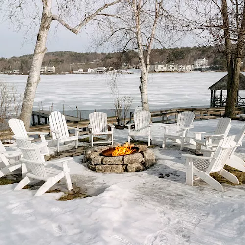 Snowy lakeside patio with white Adirondack chairs around a stone fire pit, overlooking a frozen lake and bare trees.