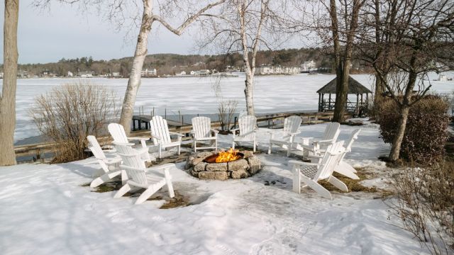 Snowy lakeside patio with white Adirondack chairs around a stone fire pit, overlooking a frozen lake and bare trees.