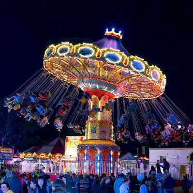 A bright nighttime carnival ride with colorful lights and a crowd gathered in front of it.