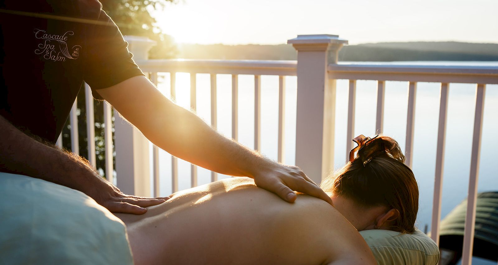 A person gives a back massage on a balcony at sunset, relaxing a guest as the ocean view glows in the background.