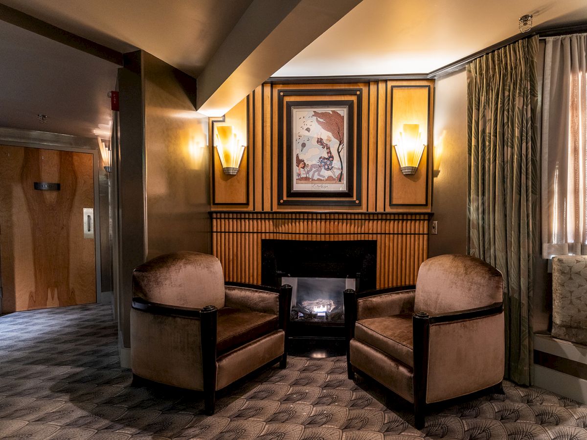 Cozy hotel lounge with two plush chairs facing a lit fireplace, framed art above, warm wall sconces, curtains, and a patterned carpet.