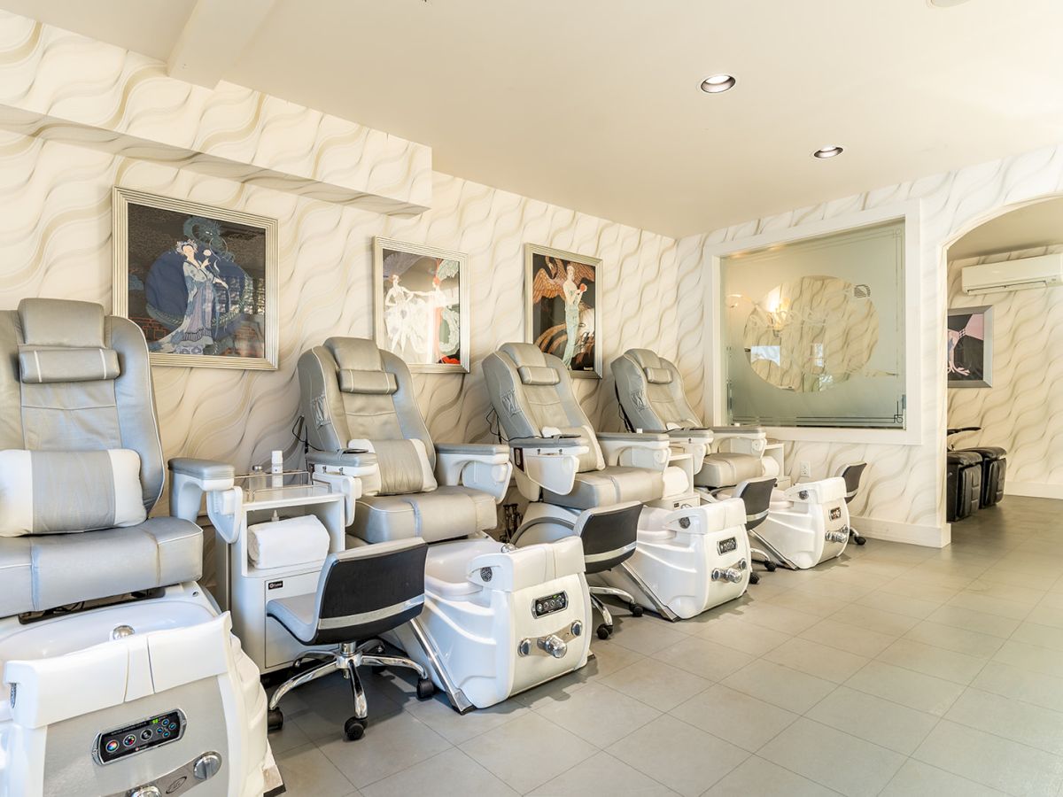 A bright modern salon with row of plush pedicure chairs, foot basins, and tech stations along a marble-patterned wall for nail spa treatments.