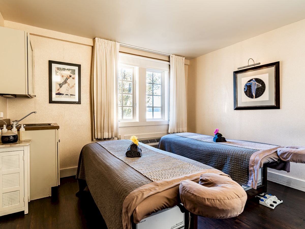 Two massage tables in a cozy, lightly decorated room with a window, framed art, and small cabinet area; calm spa vibe with soft lighting.