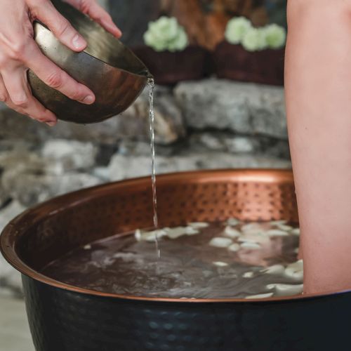 Someone pouring warm water from a small bowl into a copper tub, with a person&rsquo;s arm resting inside. Ending this sentence.
