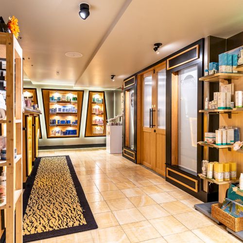 A well-lit spa or boutique hallway with wooden shelves stocked with towels, beauty products, and essentials, leading to a seating or treatment area.