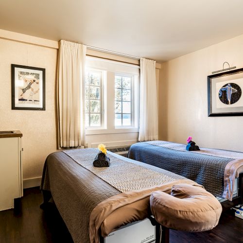 A cozy spa-like room with two massage tables, soft lighting, a window with curtains, framed art, a small cabinet, and calming decor, perfect for treatments.