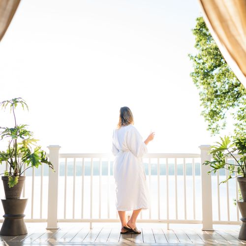 A person in a white robe stands on a bright balcony, looking out over a railing with potted plants on each side, curtains framing the view.