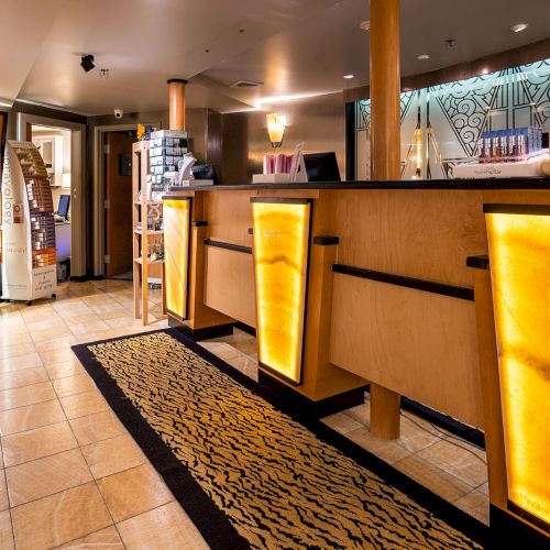 A cozy hotel lobby with a warm-lit reception desk, wooden accents, a patterned rug, and shelves of products along the left wall.