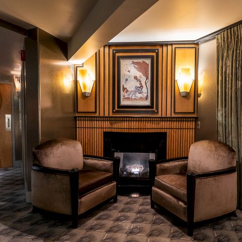 A cozy hotel lounge with two plush armchairs facing a small fireplace, framed art above, warm wall sconces, and sheer curtains by a window.