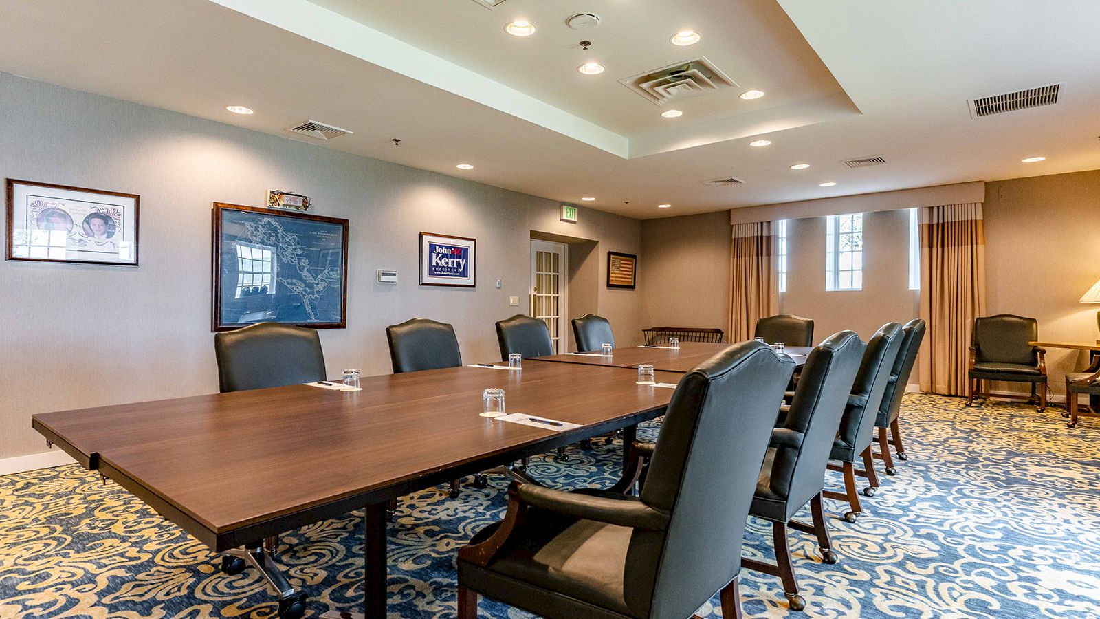 A conference room with a large table, padded chairs, framed artwork, patterned carpet, and windows with curtains.