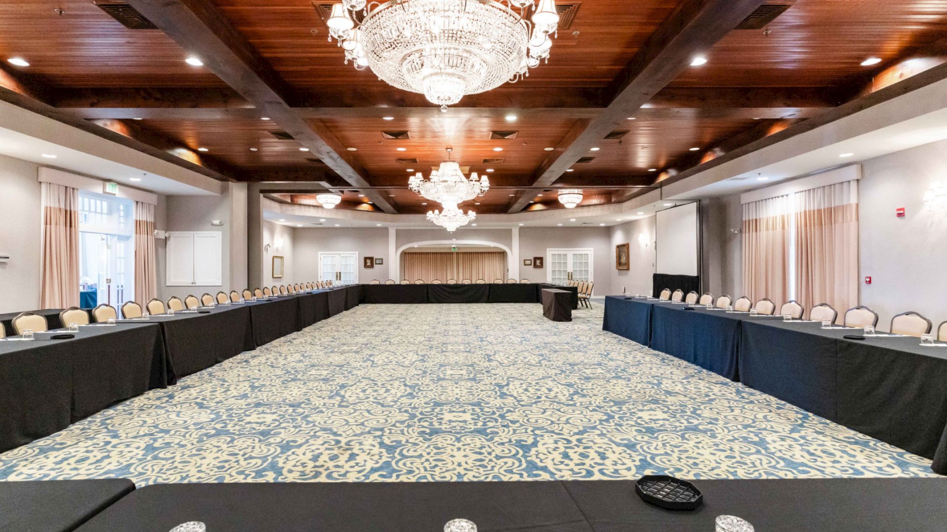 A spacious, elegant conference room with long tables arranged in a U-shape, chandeliers, and a patterned carpet, ready for a meeting or event.