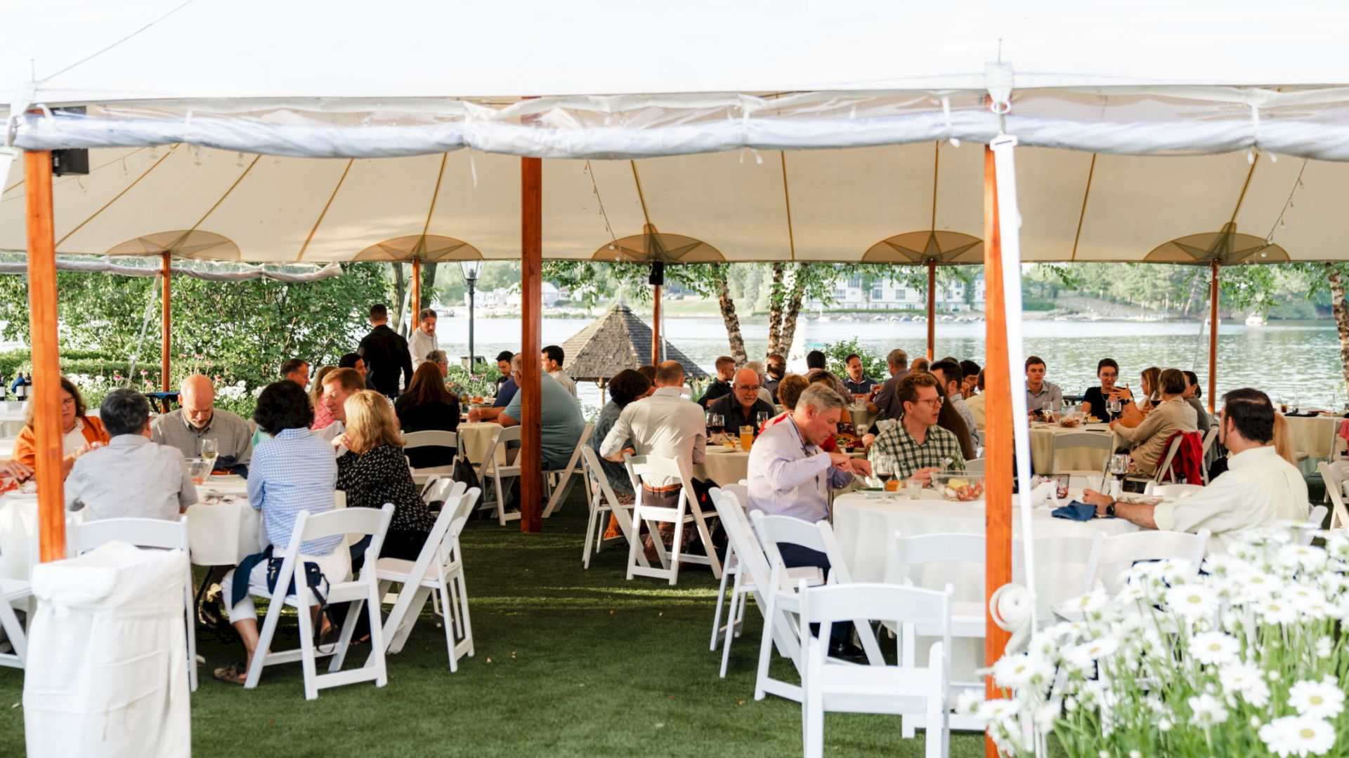 People are seated at round tables under a large outdoor tent, engaged in conversation. The setting appears to be a social or formal event.