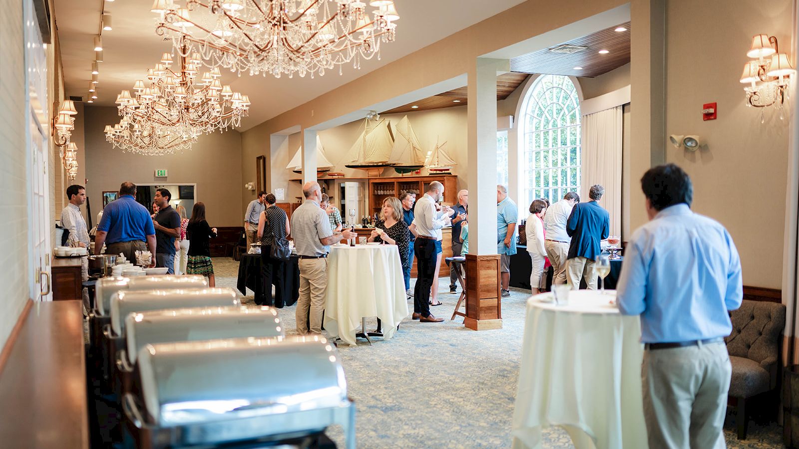 A group of people are gathered in a well-lit, elegant banquet hall with chandeliers, standing around tables, likely at a social or networking event.