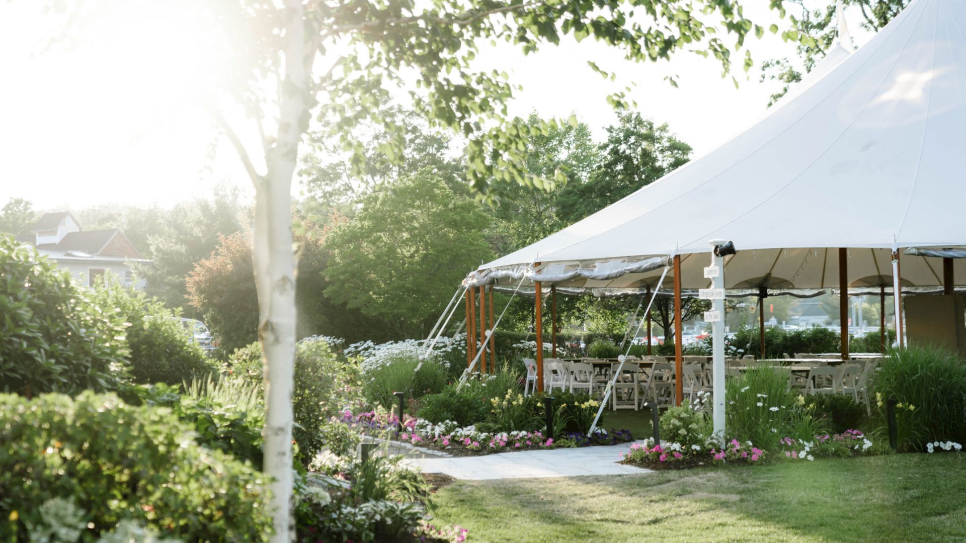 A garden scene featuring a white tent surrounded by lush greenery and flowers, with sunlight shining through the trees, creating a serene atmosphere.