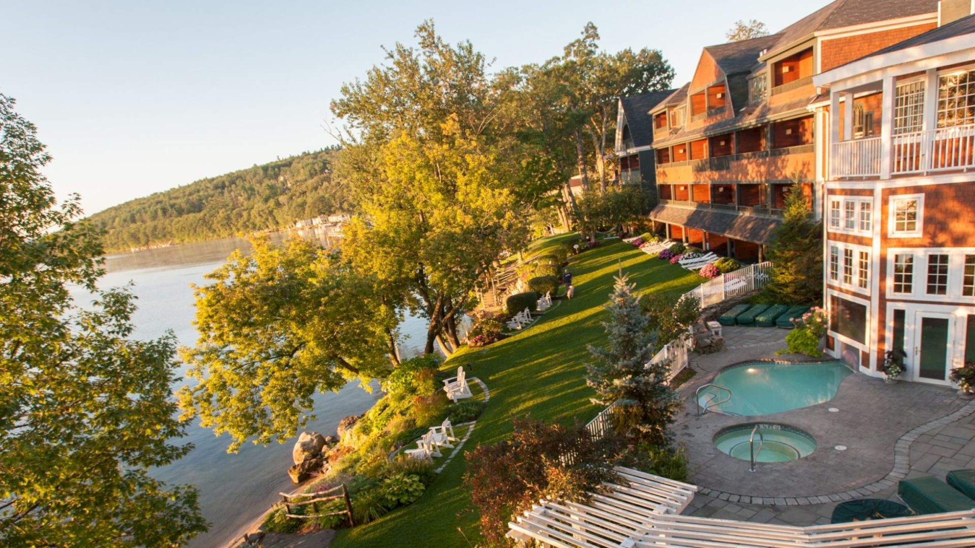 A scenic view of a lakeside resort featuring several buildings, lush greenery, a small pool, and lounge chairs, with a serene lake in the background.