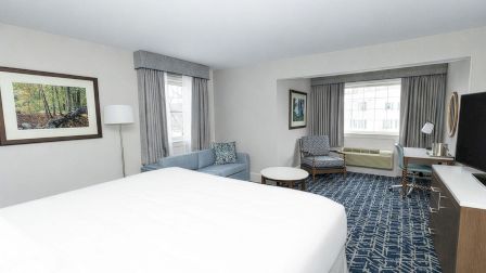 A bright hotel suite with a large bed, seating area, desk, and windows; blue patterned carpet and neutral decor.