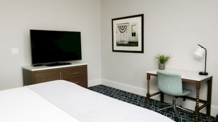 A hotel room with a bed, a flat-screen TV on a dresser, a framed picture, and a desk with a lamp, plant, and chair by the wall.