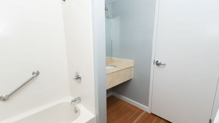 A small bathroom with a tub on the left, a sink on a counter against the back wall, and a door on the right; wood-like floor and light walls.