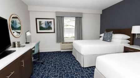A modern hotel room with two queen beds, a desk and chair, wall-mounted lamps, a flat-screen TV, a window with curtains, and neutral decor.