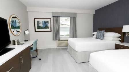 A modern hotel room with two queen beds, a desk and chair, a wall mirror, a window with curtains, and a neutral color scheme with blue accent wall.