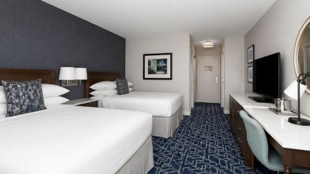 Two double beds with white linens, a dark headboard, nightstands, wall art, a desk and TV, blue carpet, and a small seating area in a hotel room.