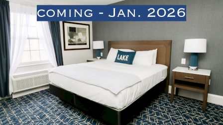 A modern hotel room with blue and white decor, featuring a bed and a "COMING - JAN. 2026" notice above.