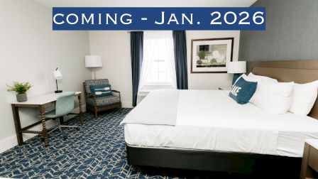 The image shows a neatly arranged hotel room with a bed, desk, chair, and a sign saying "COMING - JAN. 2026."
