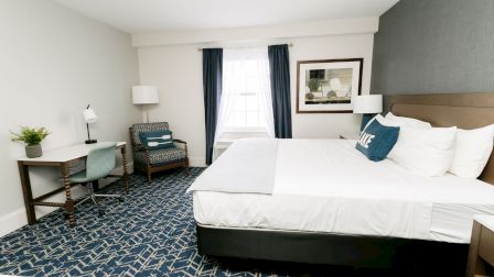 A hotel room with a large bed, nightstands, a desk and chair, a seating chair, and a window with dark blue curtains, plus wall art and a blue-patterned carpet.