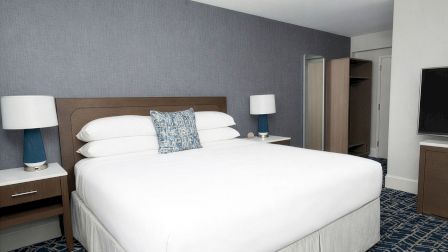 A hotel bedroom with a large white bed, two matching nightstands with lamps, a blue patterned carpet, and a flat-screen TV across from the bed, ending this sentence.