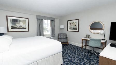 A hotel room with a large white bed, a patterned blue carpet, a window with curtains, a desk and chair, a round mirror, and a wall-mounted TV.