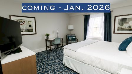The image shows a modern hotel room with a bed, TV, desk, and window. Text at the top reads "COMING - JAN. 2026."