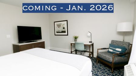 A hotel room with a bed, desk, chair, TV, and a note saying "Coming - Jan. 2026" displayed.