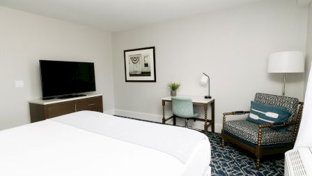 A bright hotel room with a bed in foreground, a desk with a chair, a chair and lamp, a flat-screen TV on a dresser, and a patterned carpet.