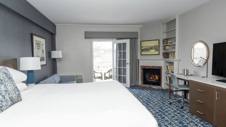 A modern hotel room with a large bed, blue patterned carpet, sitting area, desk, fireplace, and balcony door, all in neutral tones.