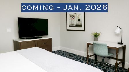 A modern hotel room with a TV, a desk, and a chair, featuring a "COMING - JAN. 2026" banner at the top of the image.