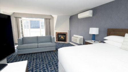 A modern hotel room with a large bed, a blue accent wall, a fireplace, a window with curtains, a gray sofa, and a ceiling air conditioner.