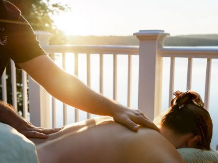 A person is receiving a back massage outdoors with a scenic view of a lake and sunset in the background.