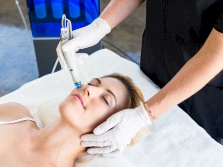 A person receives a facial treatment with a specialized device, handled by a professional wearing gloves, on a padded clinic bed.