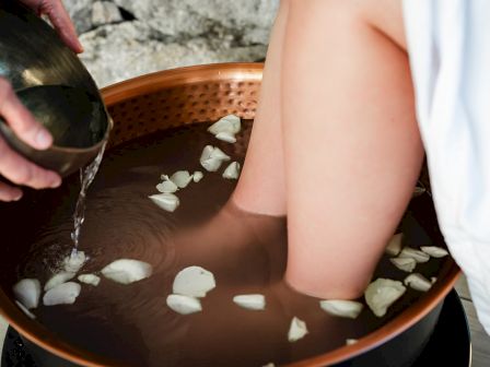 A person is soaking their feet in a foot bath filled with water and flower petals while another person pours additional water from a bowl.