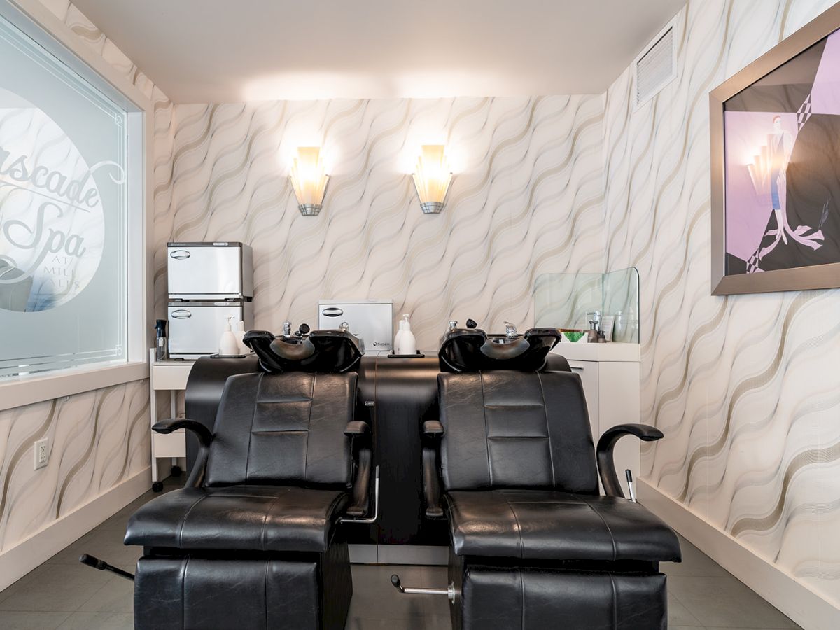A modern salon with two black reclining chairs, white marbled walls, wall lights, a small windowed area, and shampoo bowls behind the chairs, spa vibe.