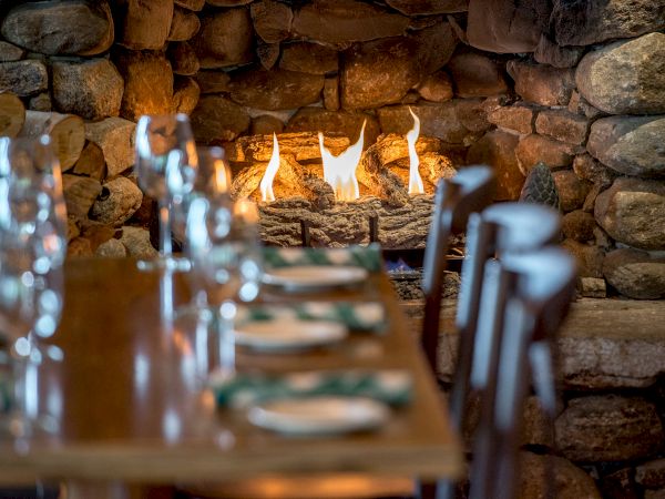 A cozy dining area with a wooden table set for a meal, in front of a lit stone fireplace giving a warm and inviting atmosphere.