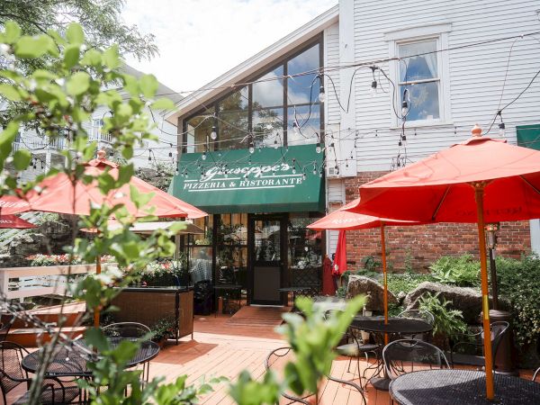 A cozy outdoor dining area with red umbrellas, a green awning entrance, and a quaint caf&eacute;/restaurant building surrounded by plants.