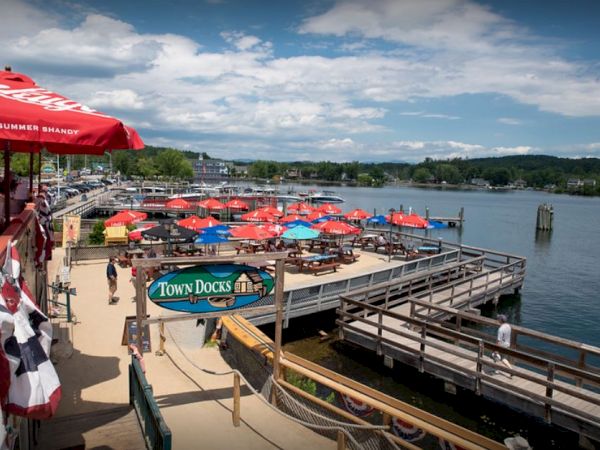 The image shows an outdoor waterside dining area with colorful umbrellas, tables, and docks, labeled "Town Docks," overlooking a scenic lake.
