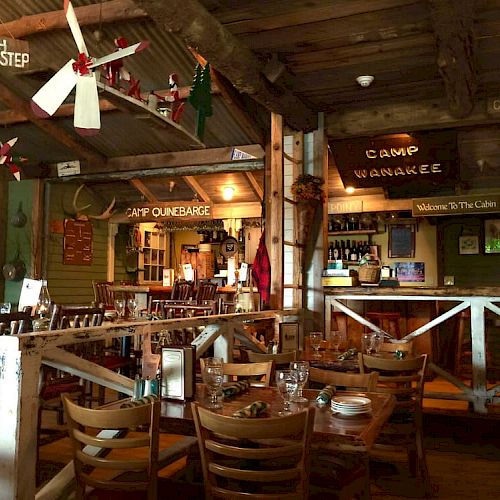 The image shows a rustic, camp-themed restaurant interior with wooden furniture, vintage decorations, and signs like "Camp Wanakee" and "Welcome to The Cabin."