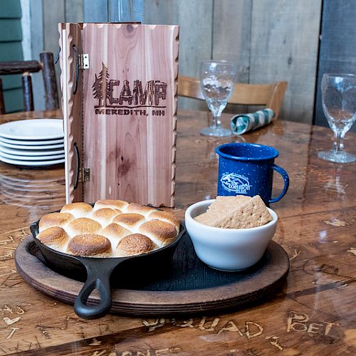 A table setting with a skillet of bread rolls, a bowl of white sauce, a blue mug, and a wooden menu displaying "CAMP Meredith, NH."