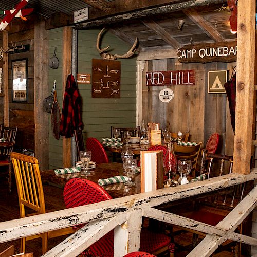 A rustic dining area with wooden furniture, antlers on the wall, checkered napkins, and signs reading "Camp Quinebaug" and "Red Hill" visible.