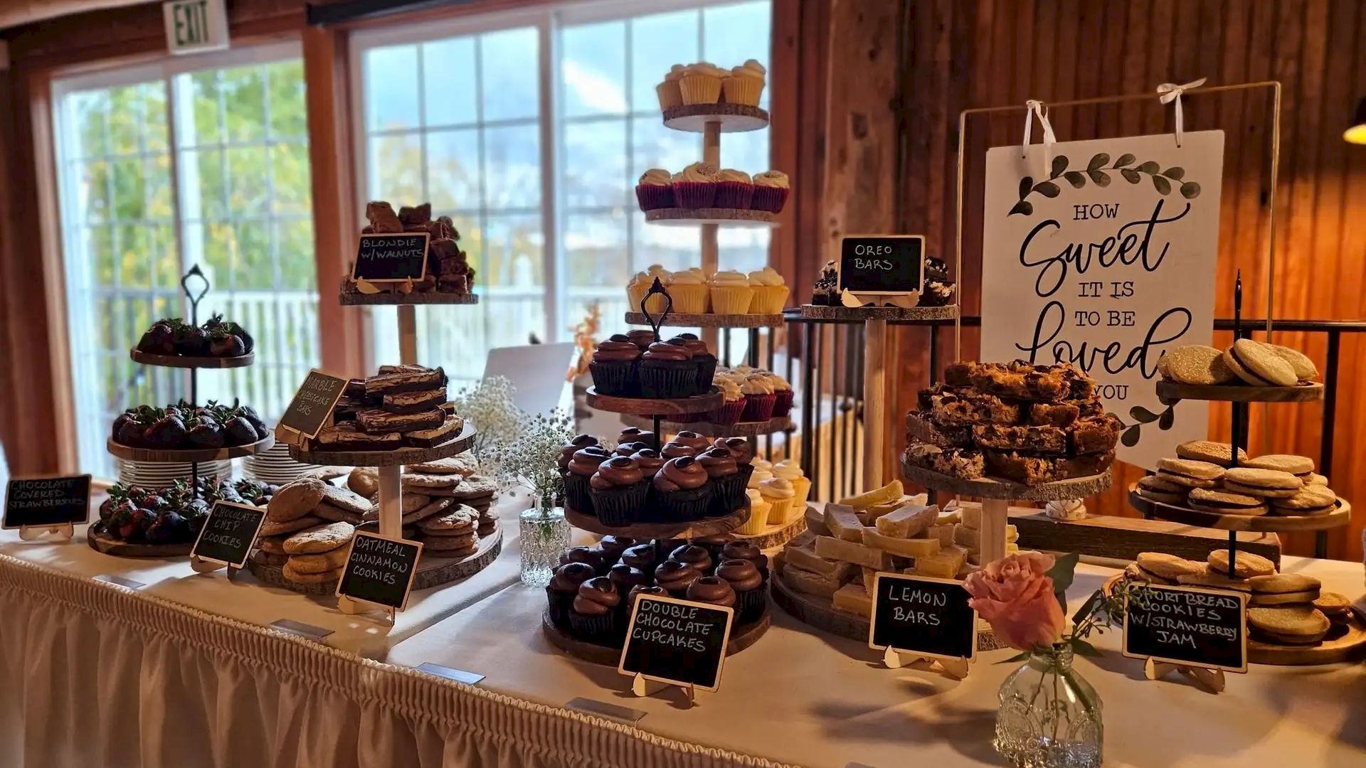 A cozy cake table with multiple stacked stands of desserts, chalkboard signs, soft lighting, and a &ldquo;You are loved&rdquo; printable sign in the background.